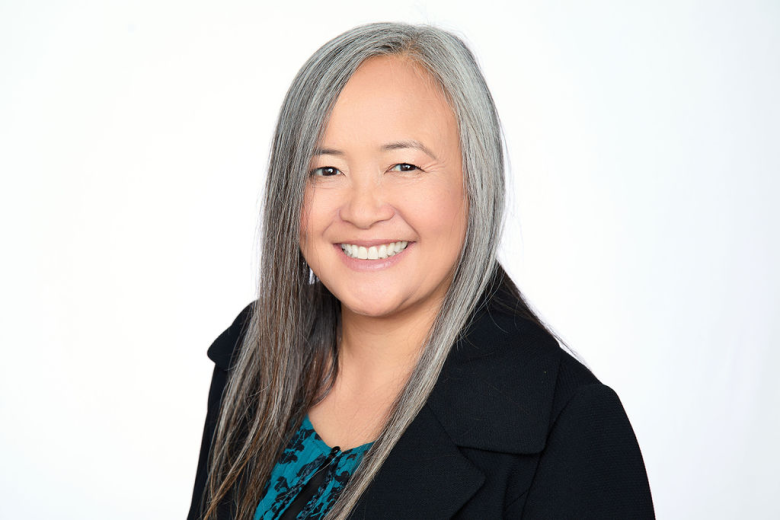 A woman with long, straight gray hair smiles at the camera in a classic Reutlinger portrait style. She is wearing a black blazer over a green top with a subtle pattern, and the background is plain white.